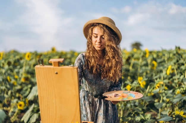 Eine junge Frau mit lockigem Haar und Hut malt in der Natur. Eine Frau steht in einem Sonnenblumenfeld an einem schönen Tag