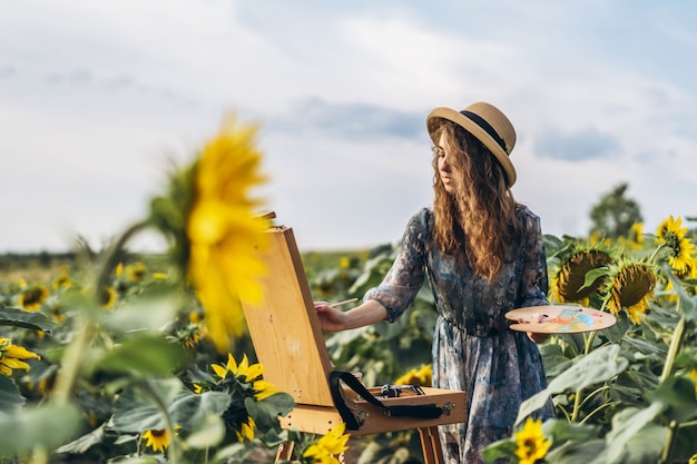 Eine junge Frau mit lockigem Haar und Hut malt in der Natur. Eine Frau steht in einem Sonnenblumenfeld an einem schönen Tag