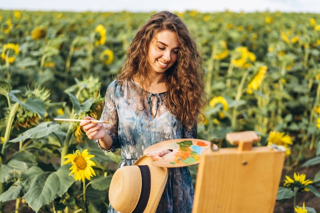 Eine junge Frau mit lockigem Haar und Hut malt in der Natur. Eine Frau steht in einem Sonnenblumenfeld an einem schönen Tag