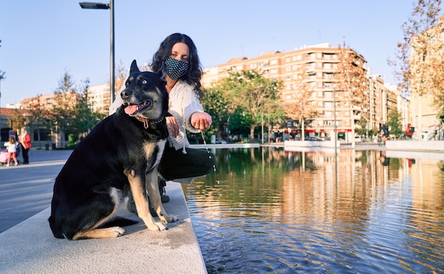 Eine junge Frau mit Maske, die in einem Parkbrunnen mit ihrem Hund sitzt. Neue Normalität. Freundschaft des Tieres mit der Frau