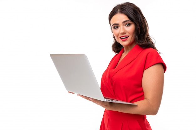 Eine junge Frau mit roten Lippen, hellem Make-up, dunklem, gewelltem, langem Haar in einem roten Anzug steht mit einem weißen Laptop da