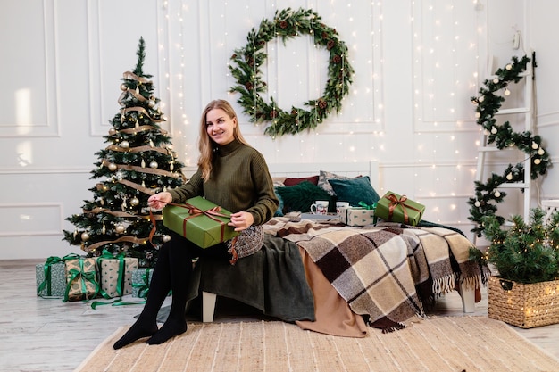 Eine junge Frau öffnet Weihnachtsgeschenke in einer Neujahrsatmosphäre und freut sich Die Frau befindet sich in einem hellen, gemütlichen Schlafzimmer, das wunderschön für Weihnachten dekoriert ist