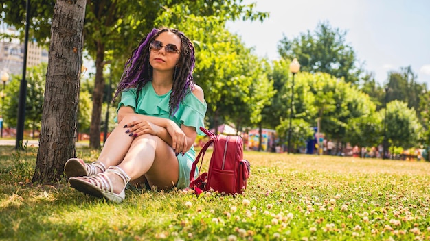 Eine junge Frau ruht sich im Schatten eines Baumes im Park aus Eine schöne junge Brünette in einem türkisfarbenen Kleid sitzt an einem sonnigen Tag auf dem Gras neben einem Baum