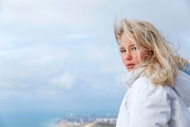 Eine junge Frau schaut auf einem Berg auf das Meer herab. Schöne Blondine in einer weißen Jacke. Aktive Erholung in der Natur Closeup