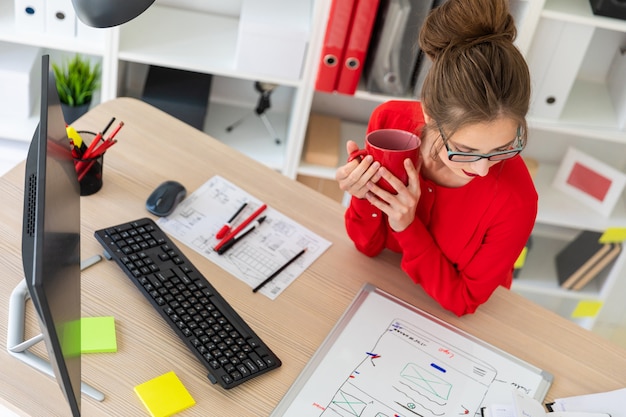 Eine junge Frau sitzt am Tisch im Büro, hält eine Tasse Kaffee und betrachtet die Magnettafel.