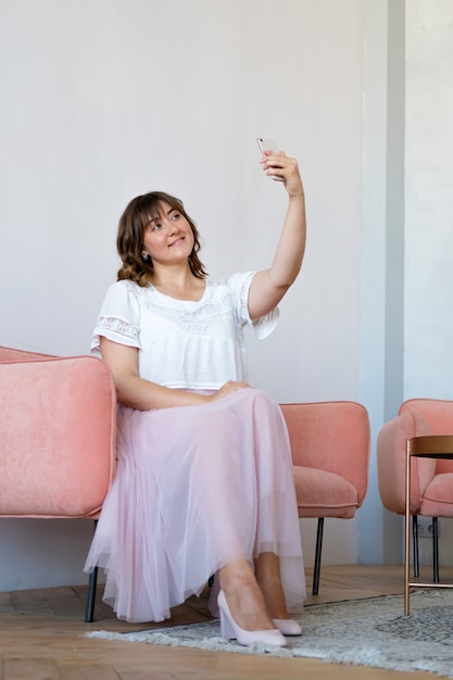 Eine junge Frau sitzt auf der Couch im Zimmer und macht ein Selfie am Telefon