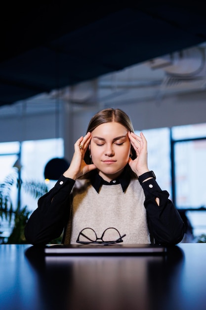 Eine junge Frau sitzt mit einem geschlossenen Laptop an einem Tisch und massiert sich den Kopf von harter Arbeit und Kopfschmerzen Frau, die an Migräne leidet