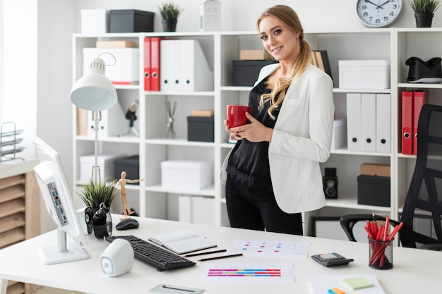 Eine junge Frau steht im Büro neben einem Computertisch und hält eine rote Tasse in den Händen.