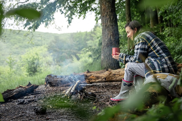 Eine junge Frau wärmt sich in der Nähe eines erloschenen Feuers mit einer Tasse wärmenden Getränks im Wald zwischen den Bäumen auf.