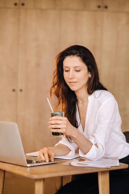 Eine junge freiberufliche Frau mit einem Cocktail in der Hand an ihrem Arbeitsplatz bei der Arbeit