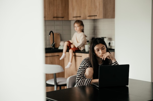 Eine junge freiberufliche Mutter in Hauskleidung, die am Laptop arbeitet und am Tisch sitzt