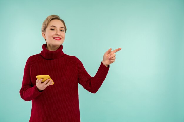 Eine junge fröhliche Frau in einem roten Pullover mit einem Smartphone auf blauem Hintergrund