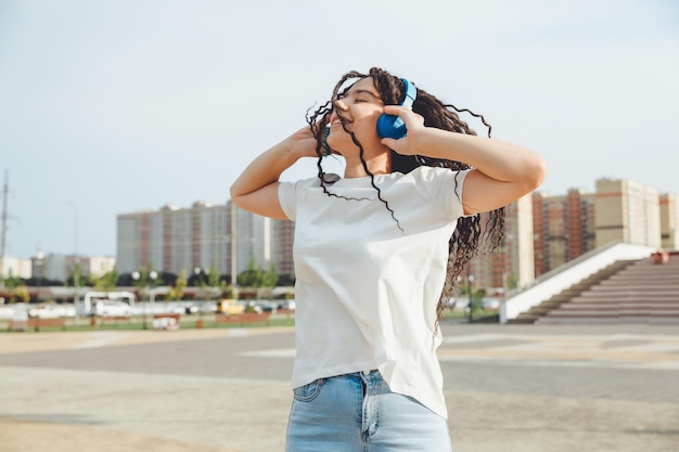 Eine junge, fröhliche, glückliche Frau mit Dreadlocks, gekleidet in ein weißes T-Shirt, tanzt und hört Musik mit Kopfhörern, die sich in einem Stadtpark entspannen und entlang einer Gasse spazieren gehen Urbanes Lifestyle-Konzept