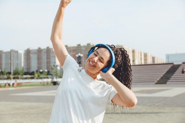 Eine junge, fröhliche, glückliche Frau mit Dreadlocks, gekleidet in ein weißes T-Shirt, tanzt und hört Musik mit Kopfhörern, die sich in einem Stadtpark entspannen und entlang einer Gasse spazieren gehen Urbanes Lifestyle-Konzept