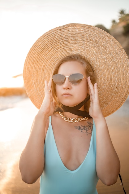 Eine junge gebräunte Frau in einem schönen Badeanzug mit Strohhut steht und ruht an einem tropischen Strand mit Sand und blickt auf den Sonnenuntergang und das Meer. Selektiver Fokus. Urlaubskonzept am Meer