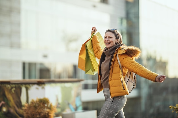 Eine junge, glücklich lächelnde Frau geht mit Einkaufstüten die Straße einer Stadt entlang.
