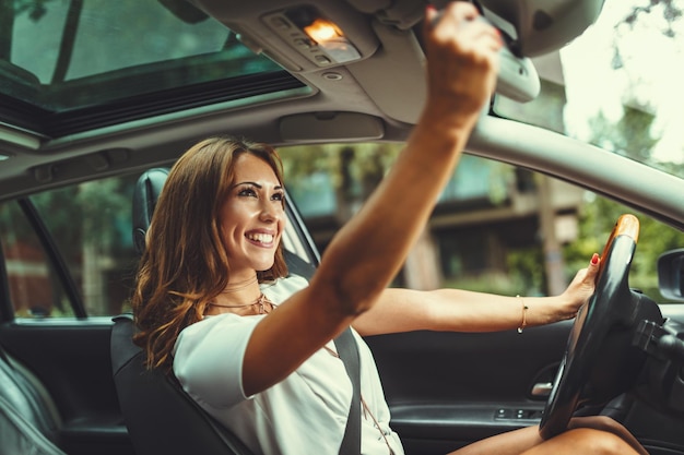 Eine junge, glücklich lächelnde Frau passt den Rückspiegel des Autos an, bevor sie ihr neues Auto fährt.