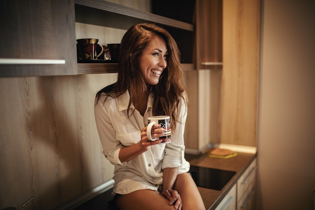 Eine junge, glücklich lächelnde Frau trinkt Morgenkaffee, während sie auf der Küchentheke sitzt.