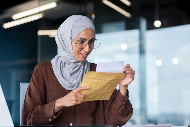 Eine junge glückliche arabische Studentin im Hijab, die am Schreibtisch im Büro sitzt und einen Umschlag hält