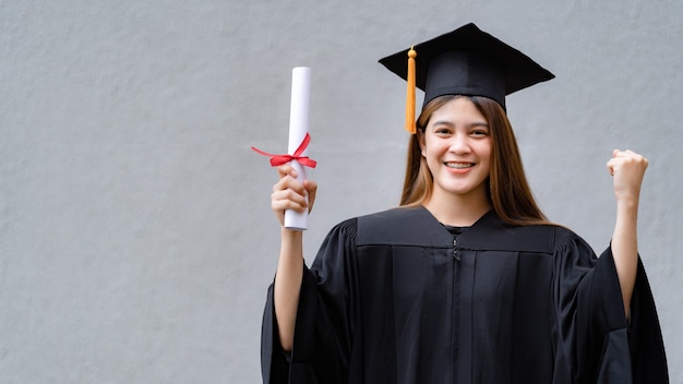 Eine junge glückliche asiatische Hochschulabsolventin in Abschlusskleid und Mörsertafel hält ein Abschlusszertifikat und feiert die Bildungsleistung auf dem Universitätscampus. Bildung stockfoto