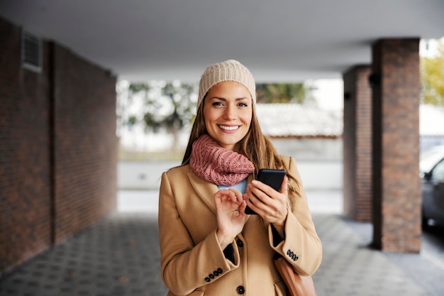 Eine junge glückliche Frau in einem warmen Outfit, die draußen steht und am Telefon tippt