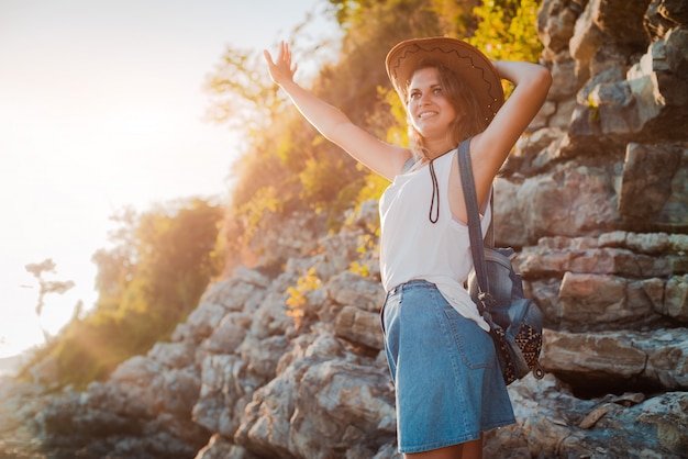 Eine junge Hippie-Frau in einem Hut und einem Rukzak mit ihren Händen oben