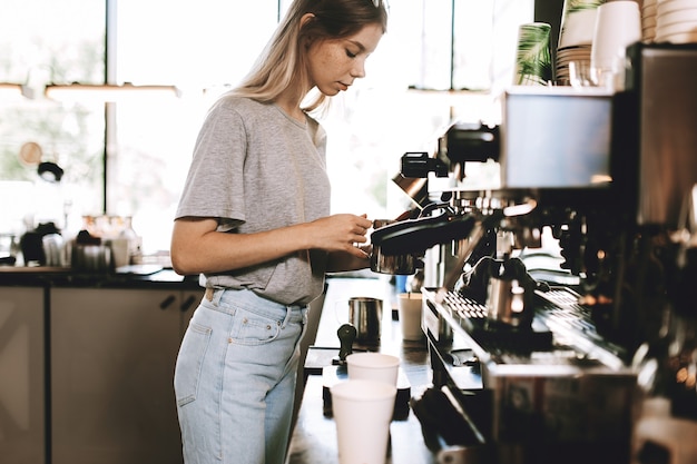 Eine junge hübsche, dünne Blondine, gekleidet in einem lässigen Outfit, kocht Kaffee in einem beliebten Café. .