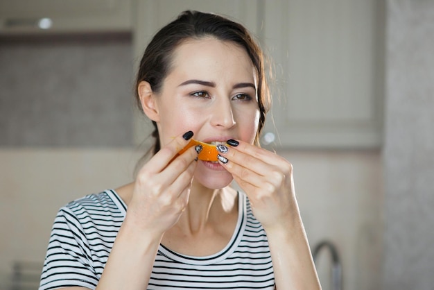 Eine junge hübsche Frau in einem gestreiften T-Shirt gießt Orangensaft in ein Glas und schneidet Obst in der Küche