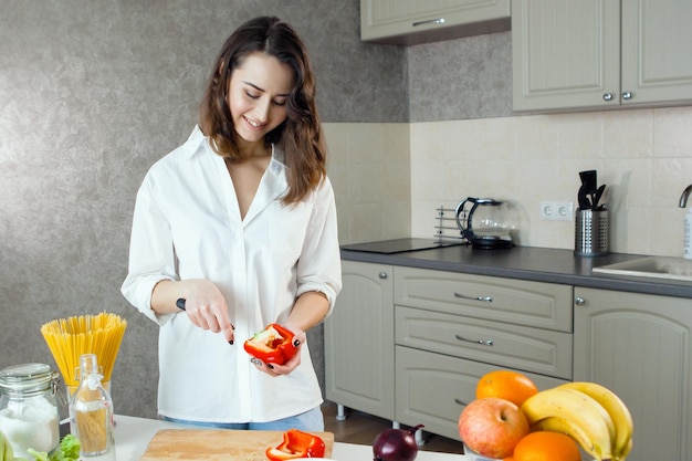 Eine junge hübsche Frau in einem weißen Hemd in der Küche schneidet Gemüse rote Paprika Mais blaue Zwiebeln Vegetarier gesunde Ernährung