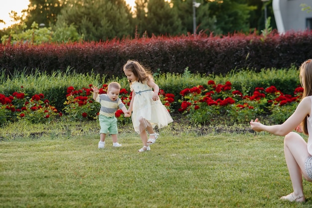 Eine junge hübsche Mutter mit ihrer Tochter hilft und bringt ihrem kleinen Sohn bei, die ersten Schritte während des Sonnenuntergangs im Park auf dem Gras zu machen.