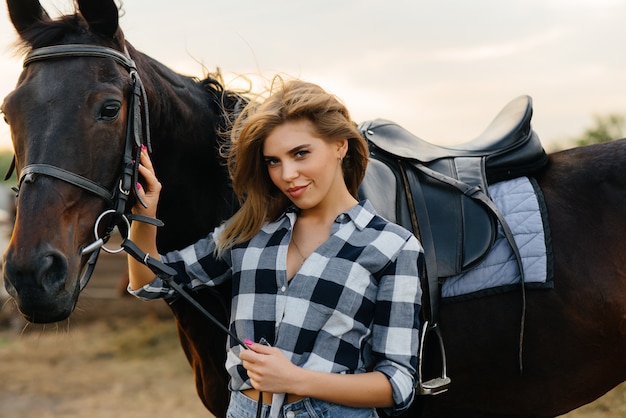 Eine junge hübsche Reiterin posiert in der Nähe eines Vollbluthengstes auf einer Ranch. Reiten, Pferderennen.