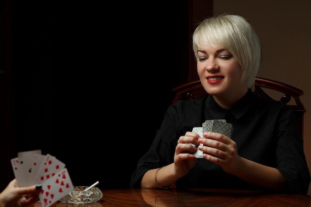 Eine junge kaukasische Blondine in schwarzer Kleidung sitzt an einem Tisch und spielt Karten mit einer anderen Frau, um sich auszuziehen und Zigaretten zu rauchen