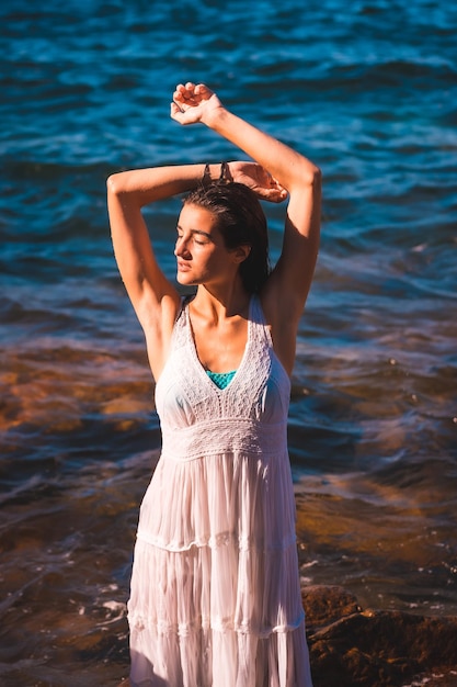 Eine junge kaukasische Frau in einem weißen Kleid und nassen Haaren sitzt auf einem Felsen am Meer