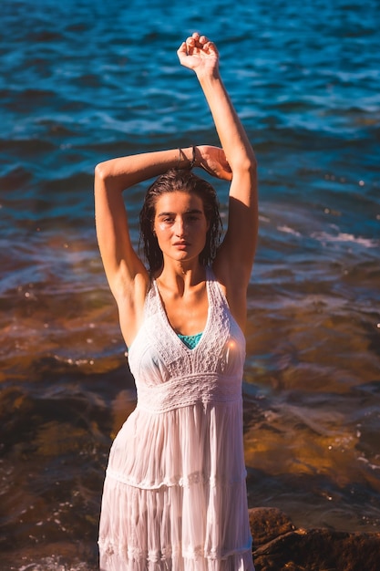 Eine junge kaukasische Frau in einem weißen Kleid und nassen Haaren sitzt auf einem Felsen am Meer