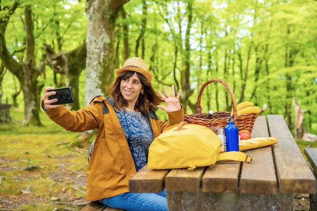 Eine junge kaukasische Mutter winkt am Telefon und sitzt an einem Picknicktisch im Wald Sommerlebensstil