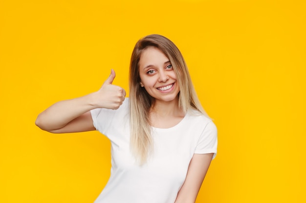 Eine junge kaukasische schöne, fröhliche, lächelnde blonde Frau in einem weißen T-Shirt, die Daumen nach oben zeigt, wie eine Geste mit ihrer Hand, die auf einer hellen gelben Wand isoliert ist?