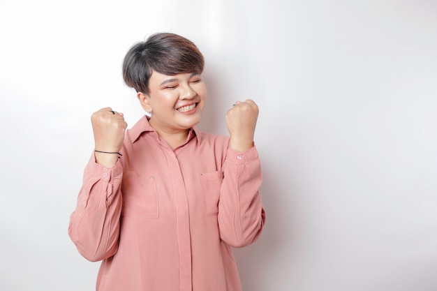 Eine junge kurzhaarige asiatische Frau mit einem glücklichen erfolgreichen Ausdruck, die ein rosafarbenes Hemd trägt, das durch einen weißen Hintergrund isoliert wird