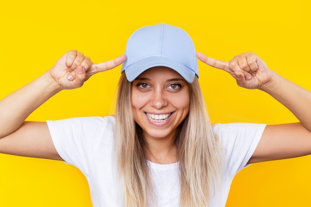 Eine junge lächelnde blonde Frau, die auf blaue Kappe zeigt, die leeren Raum auf gelbem Hintergrund zeigt