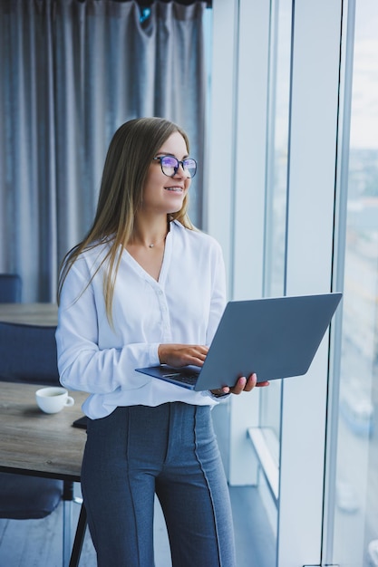Eine junge lächelnde Geschäftsfrau steht im Büro am Fenster mit einem Laptop in ihren Händen Frau-Manager in Gläsern und weißem Hemd