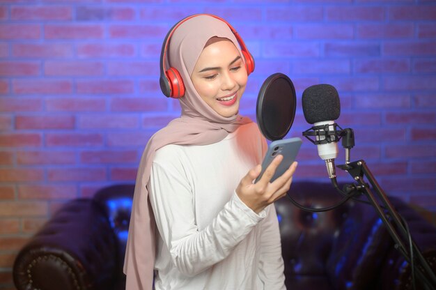 Eine junge lächelnde muslimische Sängerin, die Kopfhörer mit einem Mikrofon trägt, während sie ein Lied in einem Musikstudio mit bunten Lichtern aufnimmt.