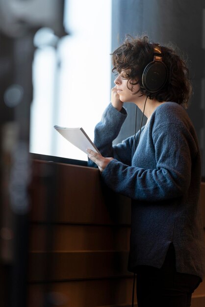 Eine junge lockige Frau schaut aus dem Fenster, hört Musik mit Kopfhörern und hält ein Notizbuch in der Hand