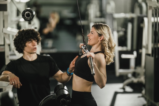 Eine junge muskulöse Frau trainiert an der Maschine, die vom Trainer im Fitnessstudio unterstützt wird.