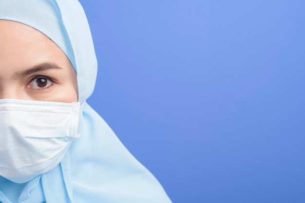 Eine junge muslimische Frau mit Hijab, die eine chirurgische Maske über Blau trägt