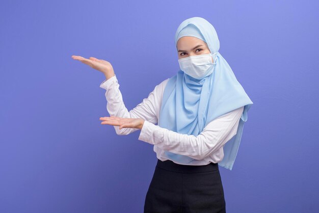 Eine junge muslimische Frau mit Hijab, die eine chirurgische Maske über blauem Hintergrundstudio trägt.