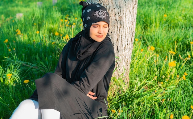 Eine junge muslimische Frau sitzt in der Natur auf dem grünen Gras im Park, ruht sich aus und lächelt