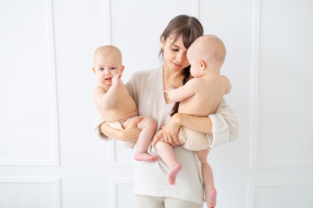 Eine junge Mutter hält zwei Zwillingsbabys in Windeln in einem hellen Raum zu Hause und lächelt glückliche Mutterschaft mit zwei Kindern