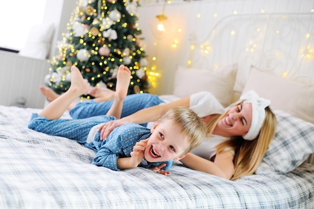 Eine junge Mutter lacht und kitzelt ihren kleinen Sohn im Pyjama, während sie auf dem Bett vor dem Hintergrund eines Weihnachtsbaums liegt. Weihnachtsfest.