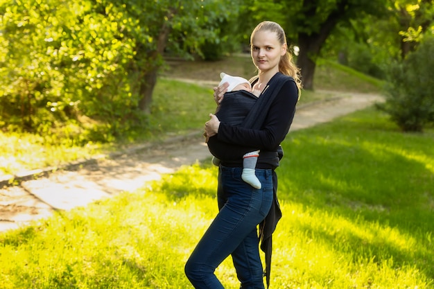 Eine junge Mutter mit ihrem Baby in einem Tragetuch steht in einem Park.