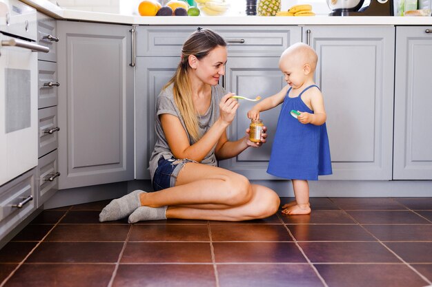 Eine junge Mutter sitzt auf dem Boden in der Küche und füttert ihre einjährige Tochter mit einem Löffel.