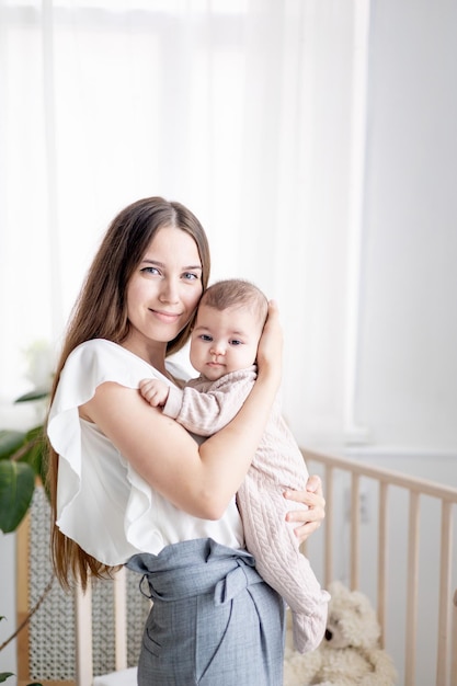 Eine junge Mutter umarmt und küsst sanft ihr kleines Mädchen, das ihn zu Hause in einem hellen Kinderzimmer neben dem Bett in den Armen hält Mütterliche Liebe und Fürsorge